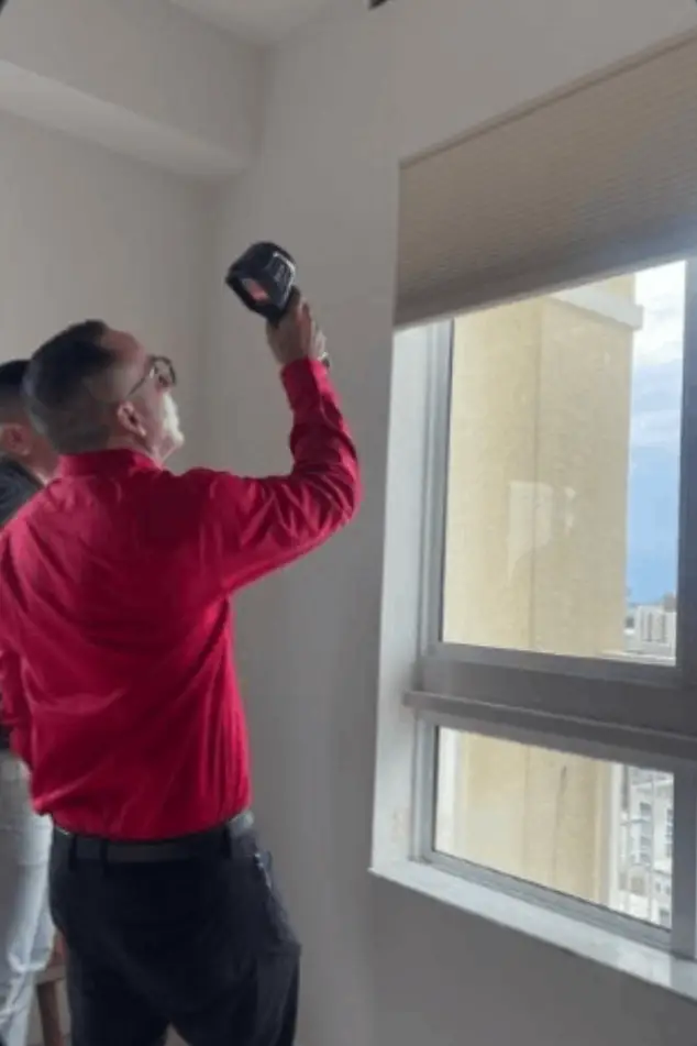 Man in red shirt using a drill on a window frame indoors.